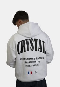 Felpa bianca con grande testo nero "CRYSTAL" sul retro, che include un indirizzo di Parigi e un dettaglio della bandiera francese. Materiale in cotone, vestibilità rilassata, design con cappuccio.