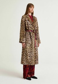 Accappatoio di lana con stampa leopardata, maniche lunghe e vita con cintura. Abbinato a pantaloni con motivo floreale e scarpe nere slip-on.