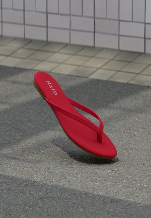 Piros tangapapucs "RAID" felirattal a talpbetéten, szürke járdán pihen egy csempézett fal szélénél.