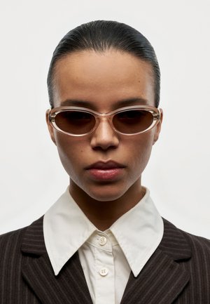 Person med glatt bakoverkammet hår iført ovale brune solbriller, en hvit knappet skjorte og en mørk blazer med stripe, vendt mot kamera.