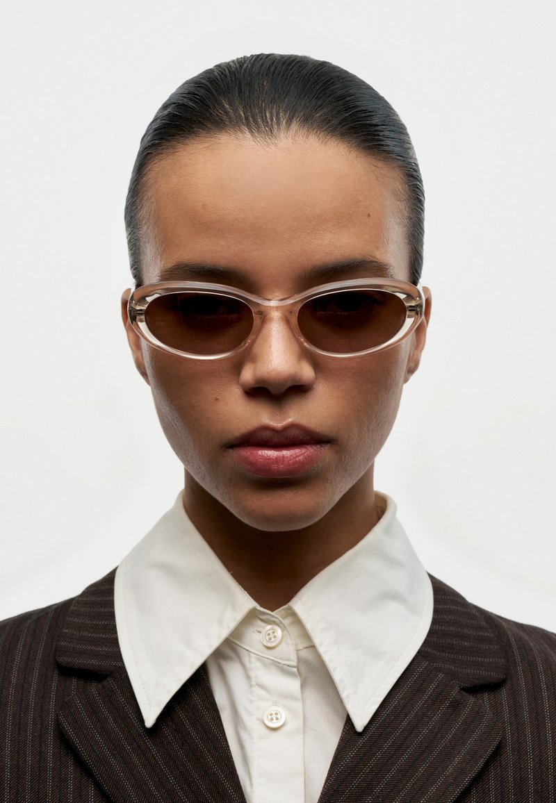 Persona con el cabello peinado hacia atrás, que lleva gafas de sol ovaladas marrones, una camisa blanca abotonada y un blazer oscuro de rayas, mirando al frente.