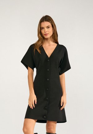 Femme portant une robe noire à manches courtes avec boutons devant et un col en V, debout devant un fond clair uni.