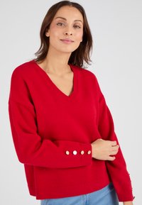 Pullover rosso lavorato a maglia con scollo a V, vestibilità morbida e spalle abbassate. Presenta bottoni in tono oro sugli orli delle maniche. Tessuto morbido.