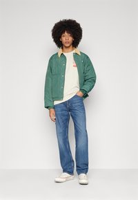 Green quilted jacket with a tan collar, worn over a light beige shirt. Paired with blue jeans and white sneakers. Casual fit and style.