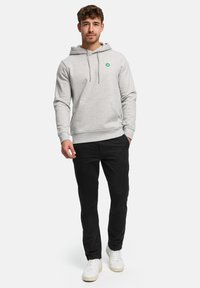 Jonge man die een lichtgrijze hoodie, zwarte broek en witte sneakers draagt, staat met één hand in zijn zak tegen een effen achtergrond.