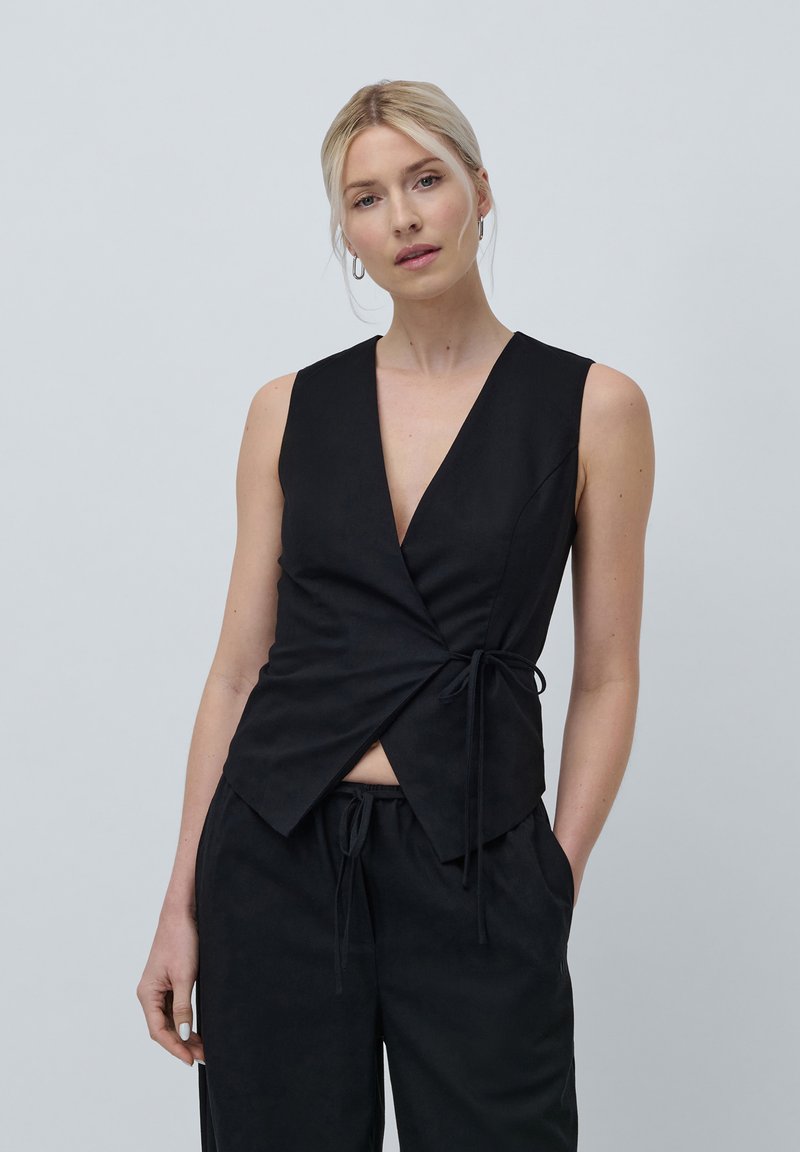 Top sin mangas negro con un profundo escote en V, detalle de lazo lateral y tejido suave. Combinado con pantalones holgados negros, creando un look coordinado.
