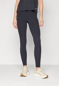 Fekete sport leggings, testhez simuló fazonban, sima textúrával és varrási részletezéssel, világos színű sportcipővel és sötét felsővel párosítva.