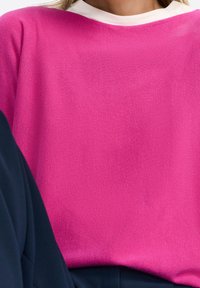 Nainen, jolla on päällään kirkkaan vaaleanpunainen neule valkoisen paidan päällä, paritettu tummansinisillä housuilla, jotka ovat lyhyet vartalon kohdalta.