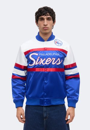 Jeune homme portant une veste Philadelphia Sixers bleue, blanche et rouge avec le logo et les inscriptions de l'équipe, debout devant un fond clair uni.