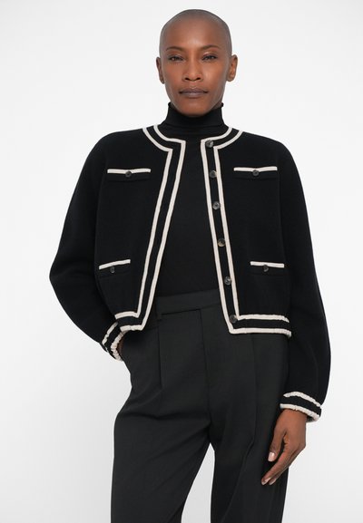 Črna kardigan s kremnimi robovi, z gumbi spredaj, dvema sprednjima žepoma in rebrastimi manšeti. Nosi se čez črn pulover s turtleneck ovratnikom in hlače.