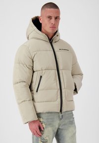 Pufferjack in lichtbeige, met een capuchon, voorkant met rits en twee zijzakken. Het materiaal heeft een gladde textuur met een gewatteerd patroon.