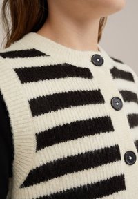 Černý a krémový pruhovaný pletený svetr s ribbovým vzorem, se čtyřmi velkými černými knoflíky na přední straně a zaobleným výstřihem.