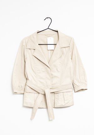 Veste beige clair ceinturée avec manches trois-quarts, col à revers, poches avant et une étiquette vierge, suspendue sur un cintre noir.
