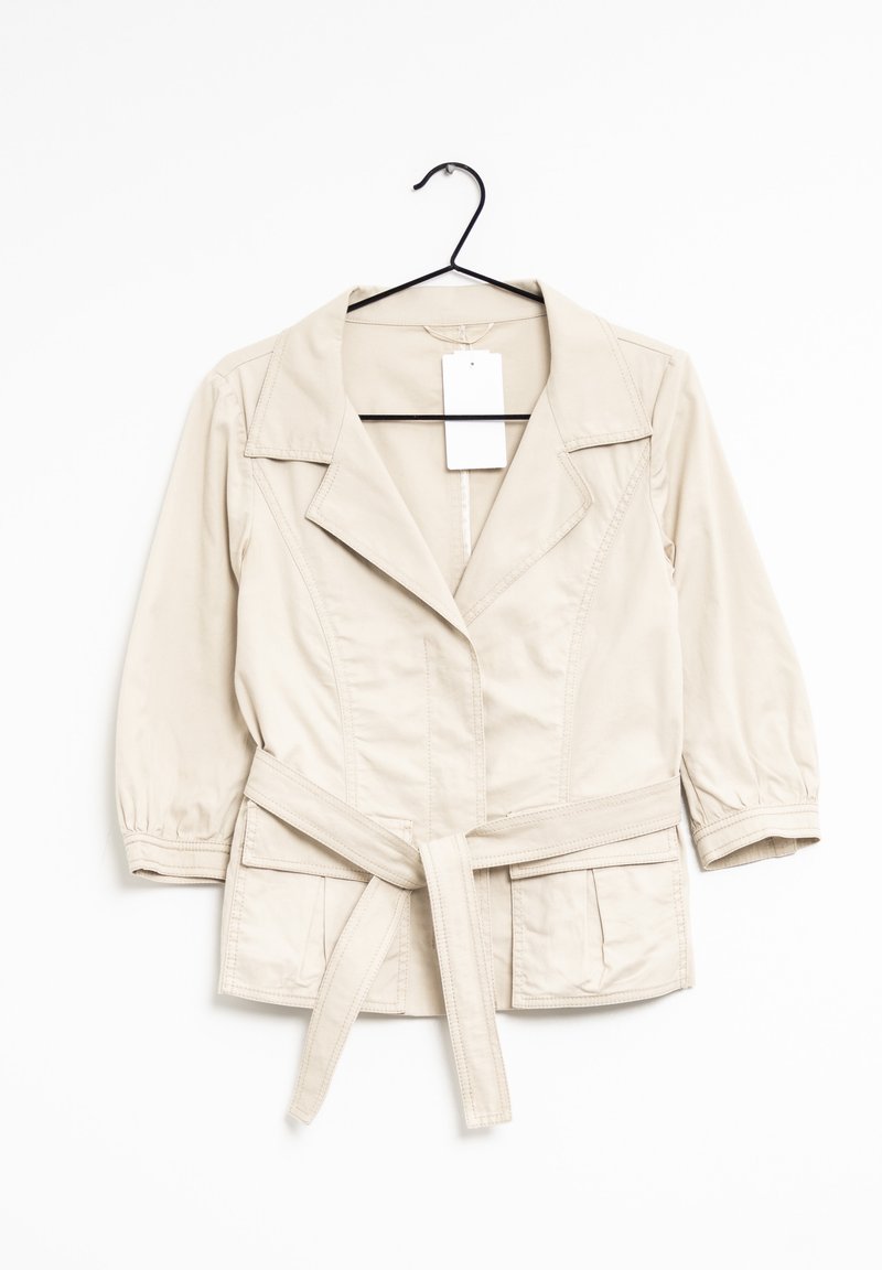 Veste beige clair ceinturée avec manches trois-quarts, col à revers, poches avant et une étiquette vierge, suspendue sur un cintre noir.