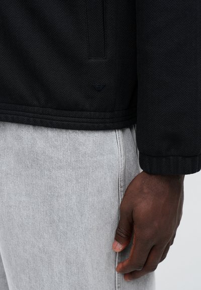 Čierny sako s rybím vzorom a hladkou textúrou, vybavené bočnou vreckom. V páre s svetlosivými džínsami.