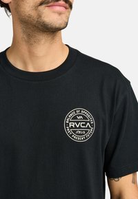 Schwarzes Baumwoll-T-Shirt mit rundem, cremefarbenem Logo auf der linken Brust. Kurze Ärmel und runder Halsausschnitt mit glatter Textur.