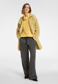 Cappotto giallo con un ampio colletto, un maglione giallo a trama e pantaloni cargo grigi a gamba larga con tasche laterali. Scarpe scure completano il look.