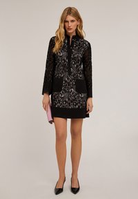 Abito in pizzo nero con maniche lunghe, chiusura con bottoni davanti e due tasche anteriori. Motivo in pizzo testurizzato con bordi neri solidi e décolleté abbinati.