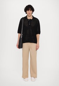 Polo en tricot noir avec une coupe ample, présentant un tissage ouvert texturé et trois boutons. Associé à un pantalon cargo beige à larges jambes, des baskets blanches et un sac noir.