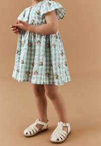 Světle modré kostkované šaty s růžovými květinovými vzory, krátkými rukávy a nařaseným pasem; spárováno s bílými sandály na pásku.