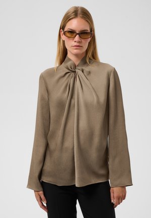 Femme aux cheveux blonds raides portant des lunettes de soleil ambre et un chemisier texturé taupe à manches longues avec un détail de lien à col montant.