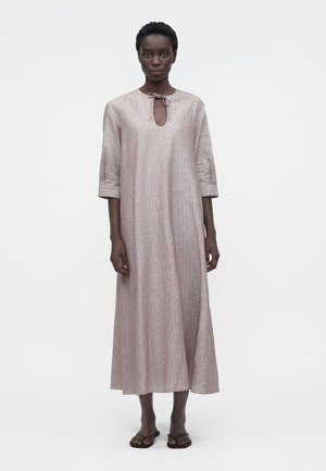 Kvinne iført en løs, ankellang, lys rosa kjole med vertikale striper, trekvart-ermer, nøkkelhull-hals med knytebånd og svarte sandaler.