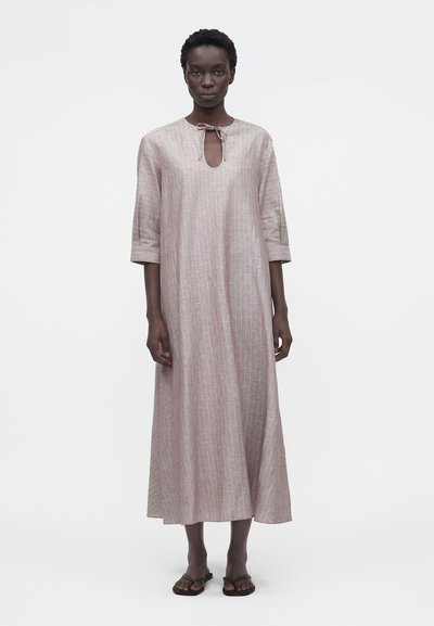 Mujer con un vestido suelto, largo hasta los tobillos, de color rosa pálido con rayas verticales, mangas tres cuartos, escote con abertura y lazo, y sandalias negras.