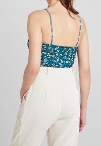 Tealfärgad blommig croptop med tunna band och rynkade detaljer, tillsammans med krämfärgade byxor med hög midja och rutigt mönster samt sidofickor.