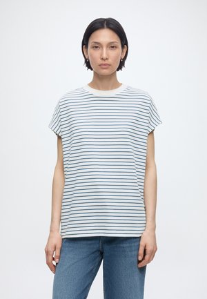 Kvinde med kort mørkt hår iført en hvid og blåstribet kortærmet top og blå jeans, stående mod en ensfarvet lys baggrund.