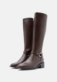 Rudos odinės iki kelių botai su lygiu paviršiumi, žemu blokuotu kulnu ir dekoratyvine juostele su apvaliu sagtimi. Užtrauktukas gale.