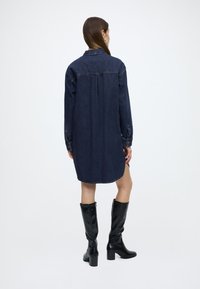 Džínové košeľové šaty v tmavomodrej farbe s golierom, dlhými rukávmi a zakriveným lemom. Kombinované s čiernymi koženými čižmami po kolená na blokovom opätku.