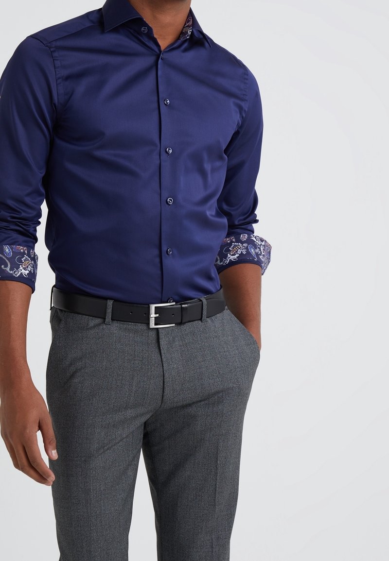 Homme portant une chemise bleu marine avec des poignets à motifs, un pantalon gris et une ceinture en cuir noir sur un fond blanc uni.
