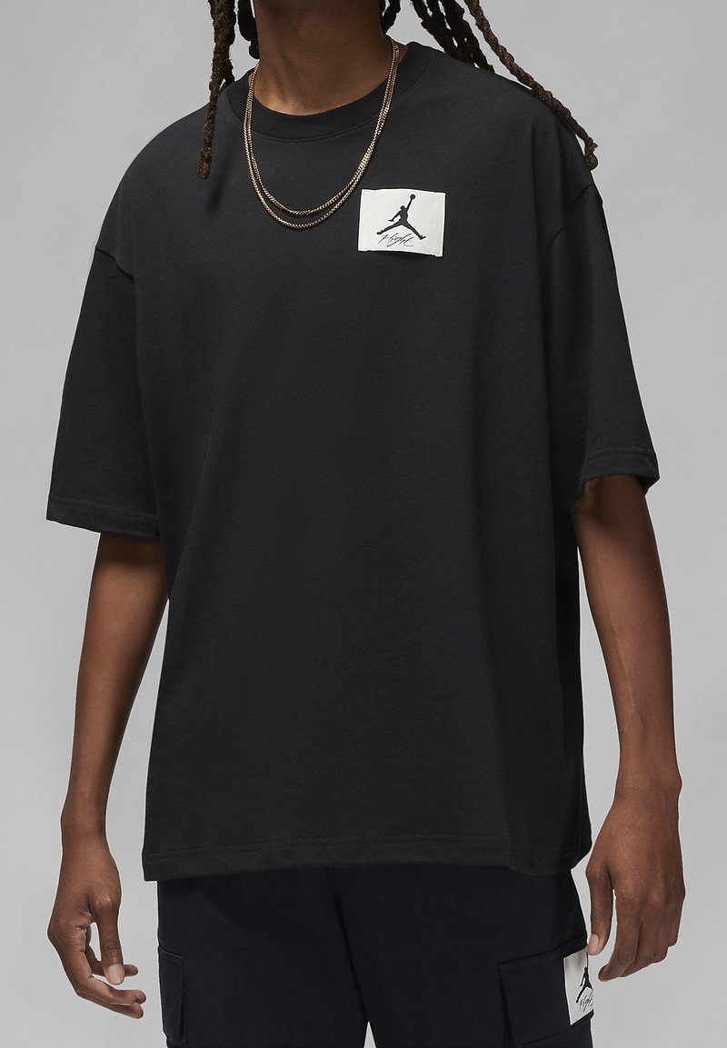 Schwarzes übergroßes T-Shirt aus Baumwolle, mit einem weißen, grafischen Logo-Patch auf der linken Brust, kurzen Ärmeln und einem lockeren Schnitt.