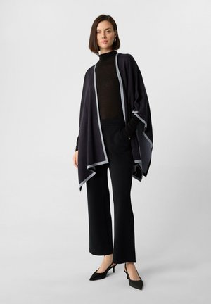 Dark gray poncho with light gray trim, featuring a flowing design. Worn over a black turtleneck and paired with black trousers and heels.