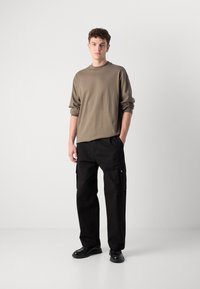 Chemise à manches longues marron clair et pantalon cargo noir avec poches latérales. Le mannequin se tient debout en chaussures noires devant un fond neutre.