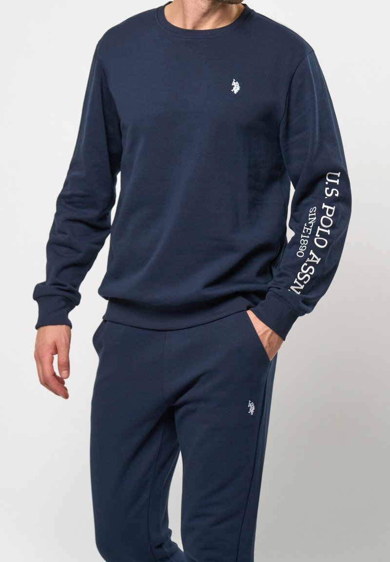 Homme portant un sweat-shirt bleu marine U.S. Polo Assn. avec le logo sur la poitrine et du texte sur la manche, pantalon assorti bleu marine avec logo sur la cuisse.