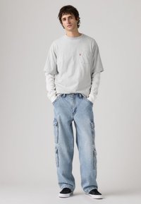 Γκρι κοντομάνικη μπλούζα πάνω από λευκό μακρυμάνικο πουκάμισο, σε συνδυασμό με ανοιχτό μπλε cargo τζιν με πλαϊνές τσέπες, φορεμένα με μαύρα αθλητικά παπούτσια.