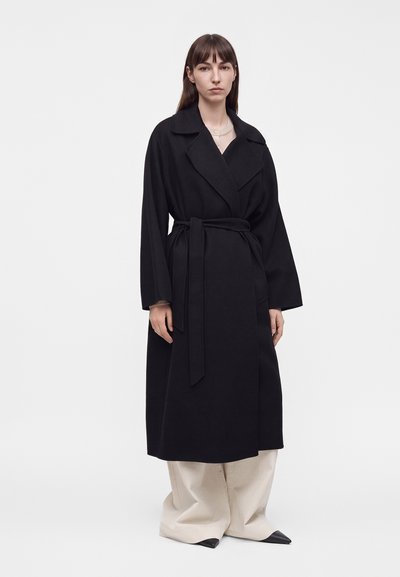 Femme debout portant un long manteau noir ceinturé sur un pantalon large beige, regardant droit devant sur un fond gris uni.