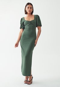 Zelena prilegajoča maxi obleka s kratkimi napihnjenimi rokavi, kvadratnim izrezom in nabiranim detajlom na prsih. Ustrezno stilizirana s črnimi sandali na peto.