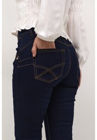 Tmavě modré džíny z denimu s kontrastním žlutým prošitím, které mají dekorativní design kapsy a slim fit. Detailní pohled ze strany.