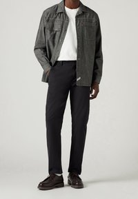 Uomo che indossa una camicia grigia scuro con bottoni sopra una t-shirt bianca, pantaloni neri e scarpe stringate marrone scuro, in piedi con una mano in tasca.