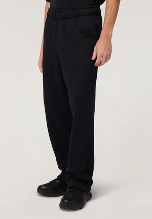 Osoba nosząca czarne spodnie dresowe z elastycznym pasem i czarne buty sportowe, stojąca na neutralnym tle.