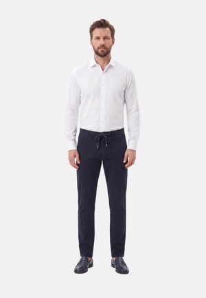 Uomo in piedi che indossa una camicia bianca abbottonata, pantaloni blu navy scuro con coulisse e mocassini neri su uno sfondo grigio chiaro.