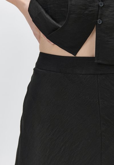 Main posée sur la taille, portant un haut noir boutonné rentré dans un pantalon noir texturé taille haute, sur un fond uni.