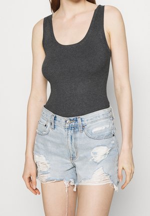 Kobieta ubrana w ciemnoszare, prążkowane, bezrękawnikowe body schowane w jasnoniebieskie, postrzępione szorty jeansowe.