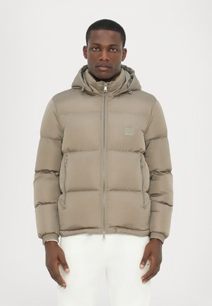 Puffer jacket in light brown, featuring a hood, zip front, and stitched padding. Includes side zip pockets and logo accent on chest.
