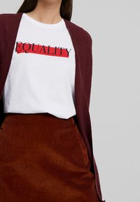 Vitt T-shirt med svart och röd texten "EQUALITY", lager på lager under en vinröd ribbad cardigan, parat med en brun kordflodbyxa.