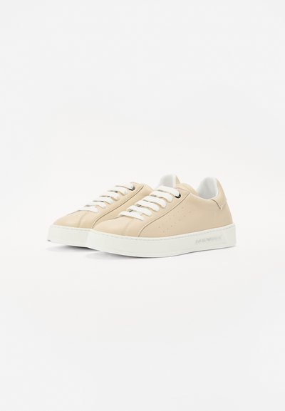 Beige leather sneakers with white rubber soles, featuring perforated details, white laces, and a smooth texture. Minimalist design.