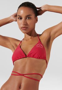 Top bikini rosso a triangolo con spalline a fascia e sottogola arricciato. Presenta una stampa del logo bianco sul lato sinistro e dettagli con cinghie incrociate sul retro.