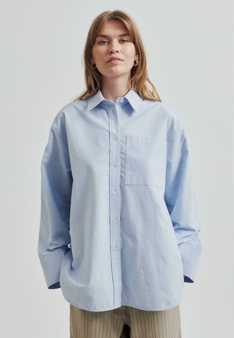 Cămașă oversized de culoare albastru deschis, din bumbac, cu un singur buzunar pe piept, mâneci lungi și design clasic al gulerului.